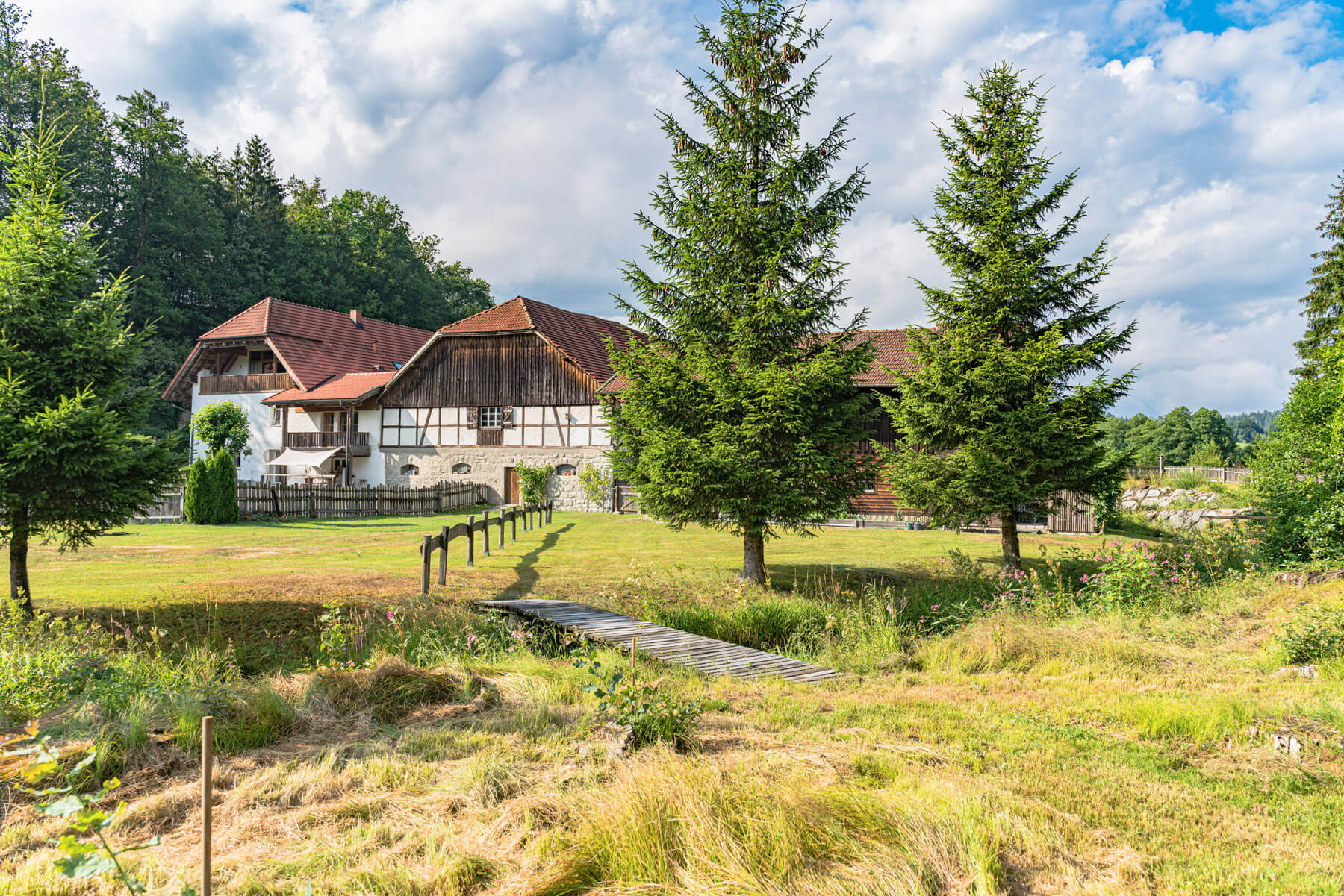 Hinter grünen Rasenflächen, hohen immergrünen Bäumen und einer kleinen Holzbrücke steht unter einem teilweise bewölkten Himmel ein rustikales Bauernhaus mit roten Dächern und Holzwänden.