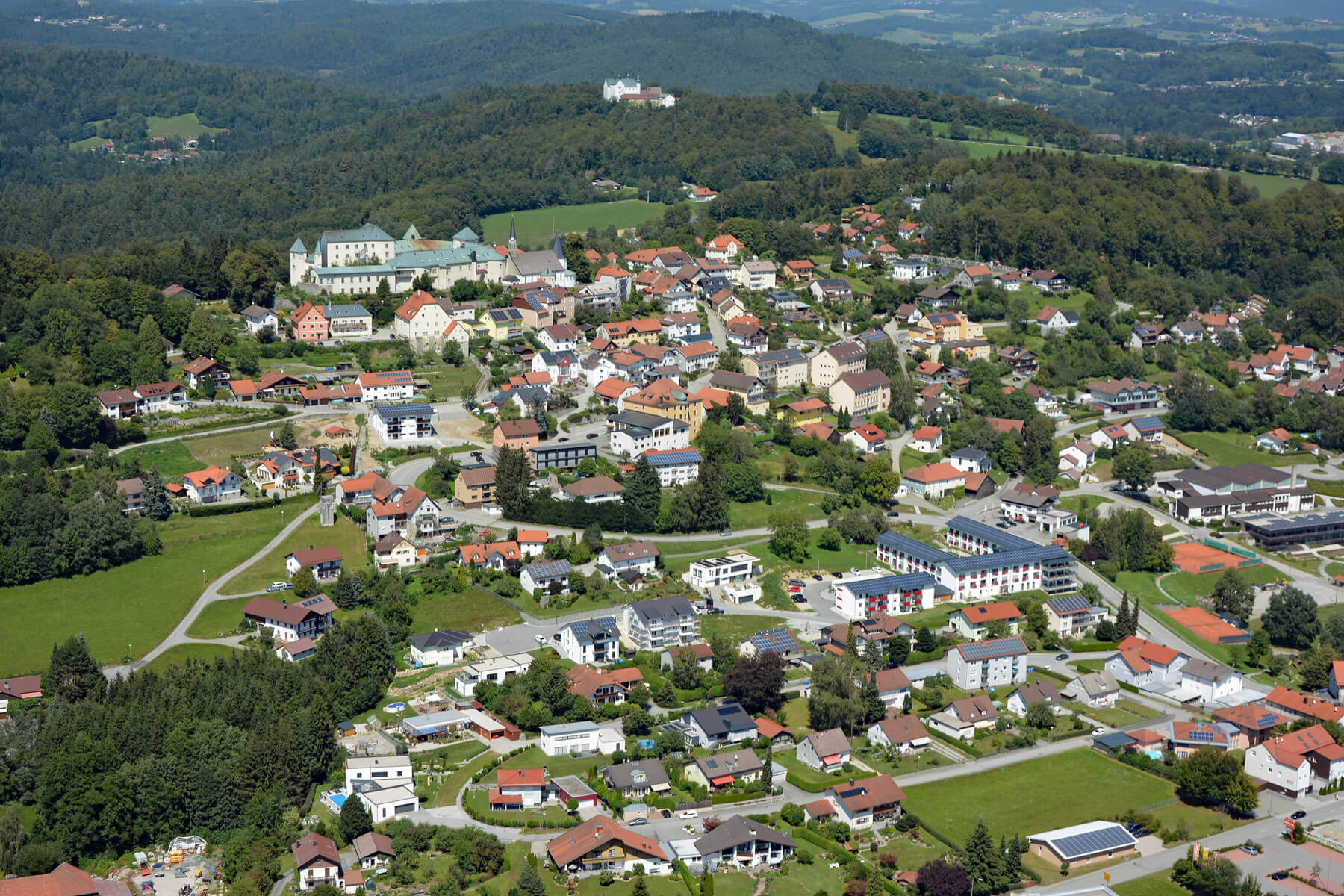 Luftaufnahme eines europäischen Dorfes mit Häusern mit roten Dächern, größeren Gebäuden, gewundenen Straßen, grünen Feldern und einem bewaldeten Hügel im Hintergrund, auf dem sich ein großes Gebäude sowie ein weißes Bauwerk in der Ferne befinden.