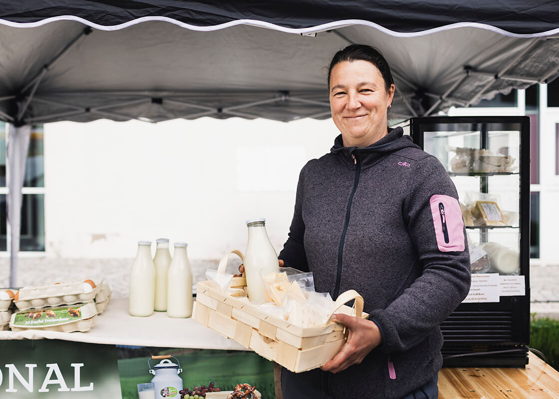 Eine Frau steht an einem Marktstand, lächelt und hält einen Korb mit einer Milchflasche aus Glas und Käse in der Hand. Auf einem Tisch neben ihr sind Milchflaschen und Eierkartons ausgestellt. Im Hintergrund steht unter einem Vordach eine Kühlvitrine.
