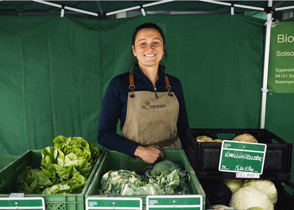Eine Frau in einer Schürze steht hinter Kisten mit frischem Gemüse und lächelt an einem Marktstand mit grünen Tischdecken.