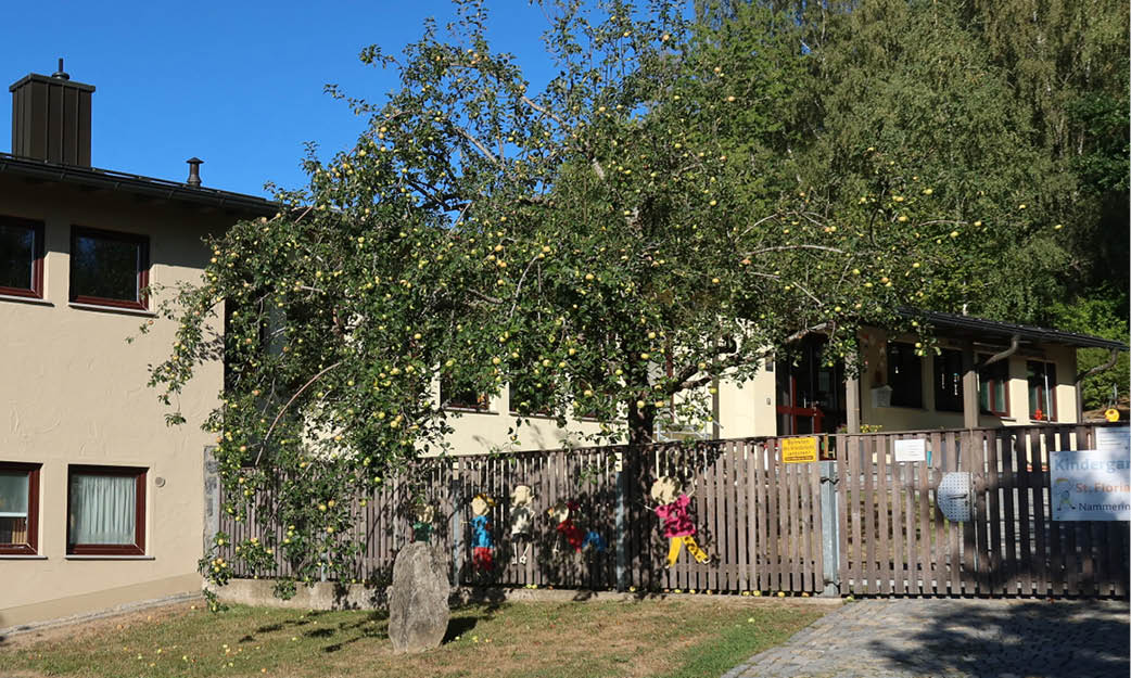 Vor einem beigen Gebäude mit braunen Zierleisten steht hinter einem Holzzaun ein großer Baum mit grünen und gelben Früchten. Am Zaun hängen bunte Kinderdekorationen. Im Vordergrund sind Gras und Kopfsteinpflaster zu sehen.