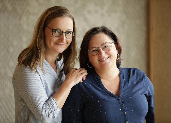 Zwei Frauen mit Brille und langen Haaren stehen dicht beieinander und lächeln in die Kamera. Die Frau links legt ihre Hand auf die Schulter der anderen. Sie befinden sich in einem Innenraum vor einer neutralen, strukturierten Wand.